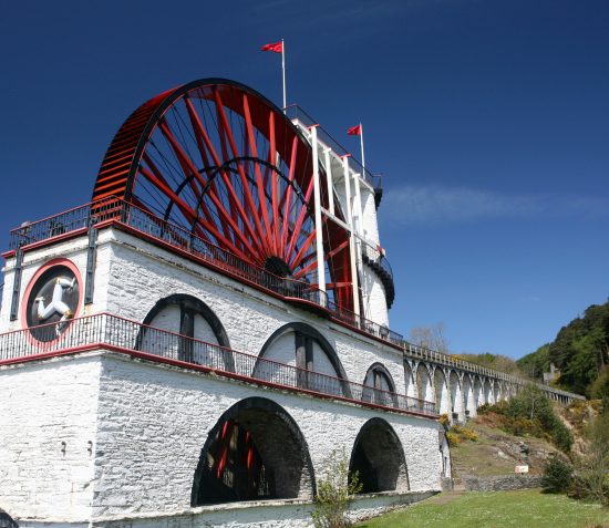 Wide angle colour photograph of the Laxey Wheel on a sunny day with green trees in the background